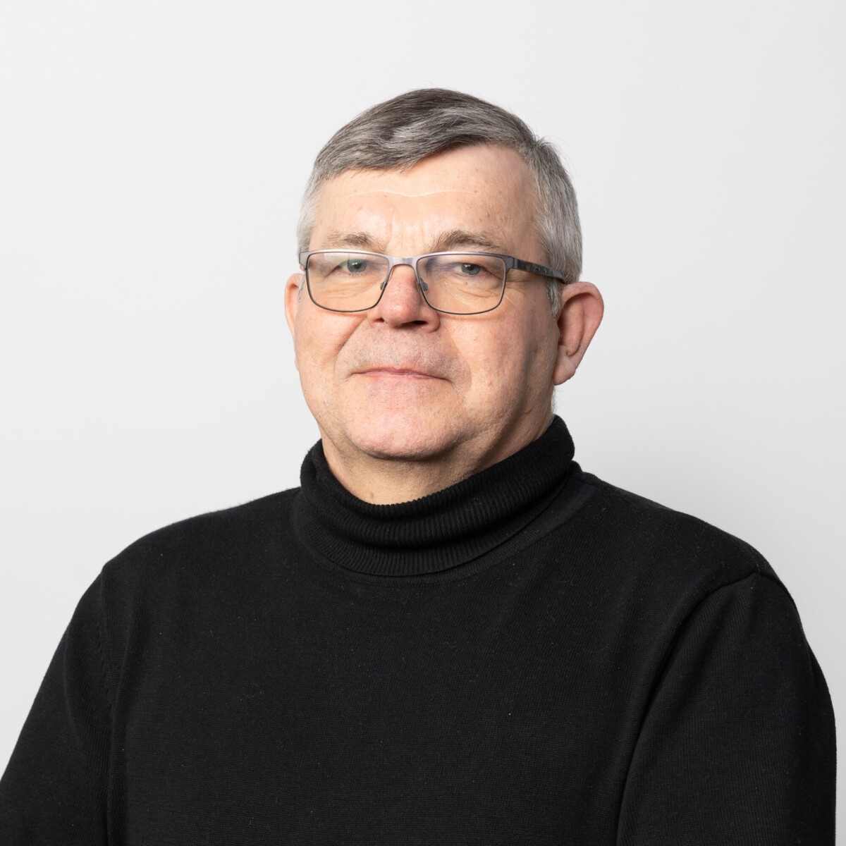 Keski-ikäinen mies, jolla on lyhyet harmaat hiukset ja silmälasit, on pukeutunut mustaan poolopuseroon ja hymyilee pehmeästi, poseeraa valkoista taustaa vasten.