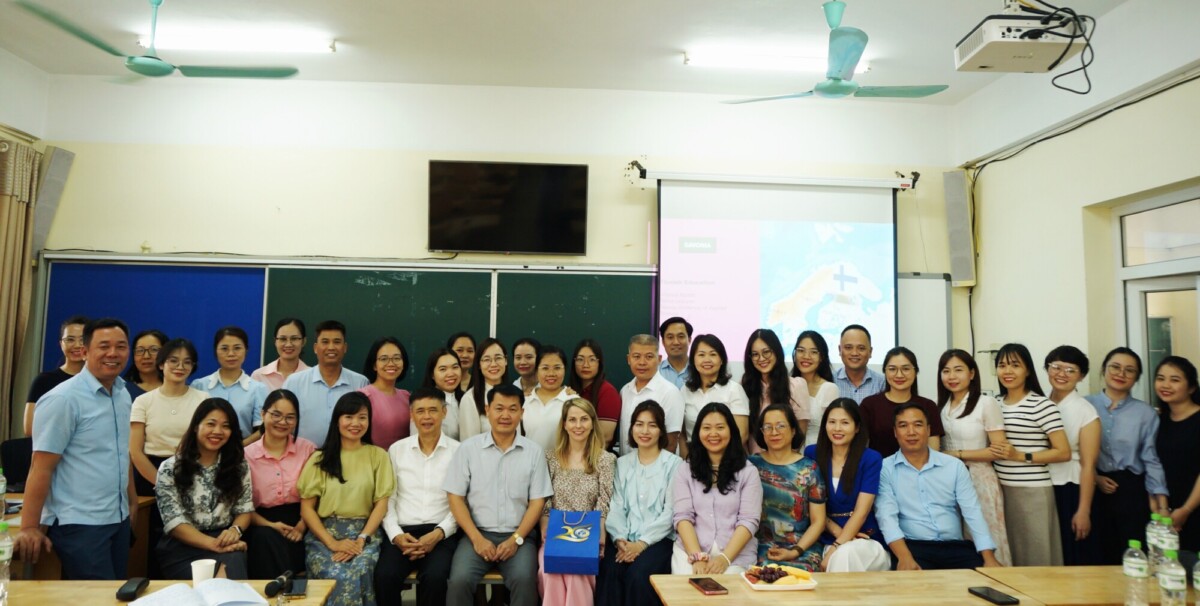 Suuri joukko ihmisiä, sekä miehiä että naisia, poseeraa ja hymyilee yhdessä luokkahuoneessa, jonka taustalla näkyy taulu, projektorinäyttö ja kattotuulettimet.