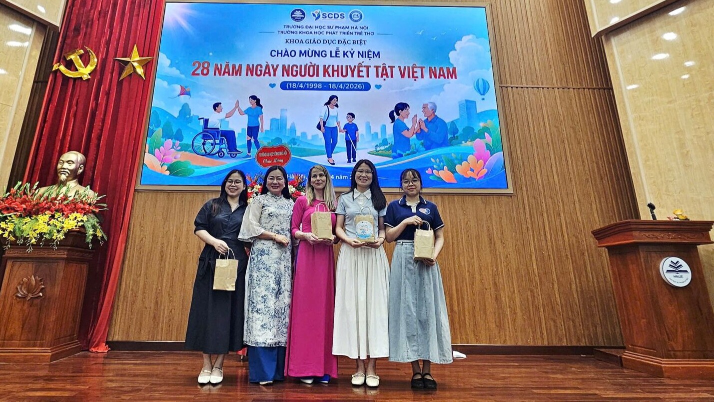 Viisi naista seisoo lavalla lahjapussit kädessään ja hymyilee valokuvaan. Heidän takanaan on suuri värikäs banderolli, jolla juhlistetaan Vietnamin 28. vammaisten päivää. Lavalla on koroke, kukkia ja puupanelointi.