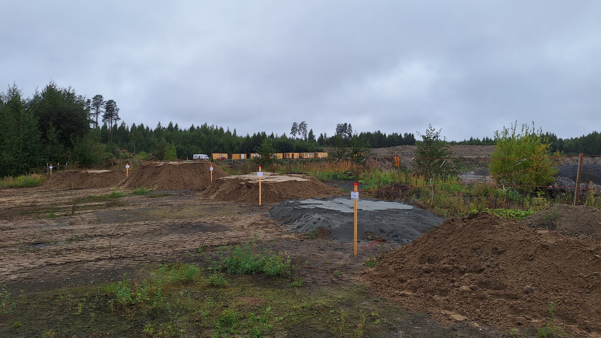 Samea rakennustyömaa, jossa on maakasoja, puukeppejä, jotka merkitsevät alueita, niukkaa kasvillisuutta ja puita pilvisen taivaan alla.