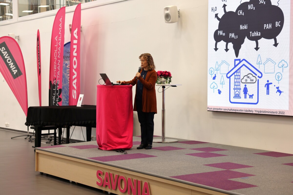 Nainen seisoo korokkeella, jossa lukee SAVONIA, ja pitää esitelmää, jossa suurella näytöllä näkyy kaavio ilmansaasteista ja taloista. Taustalla näkyy vaaleanpunaisia lippuja ja banderolleja, joissa on sana SAVONIA.