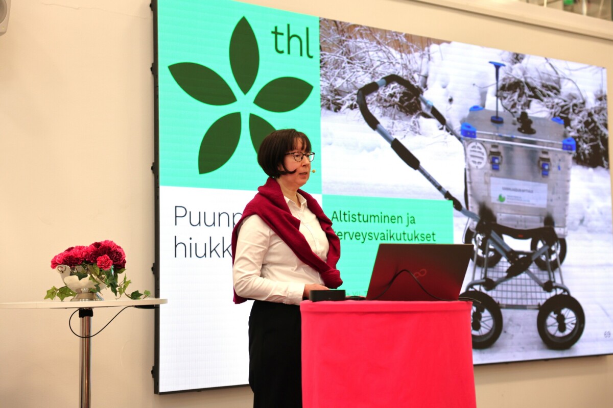 Nainen seisoo korokkeella pitämässä esitelmää kannettavan tietokoneen kanssa. Hänen takanaan on näyttö, jossa näkyy THL:n logo, suomenkielistä tekstiä ja kuva rattaisiin kiinnitetystä laitteesta lumisessa ympäristössä. Lähistöllä pienellä pöydällä on kukkamaljakko.