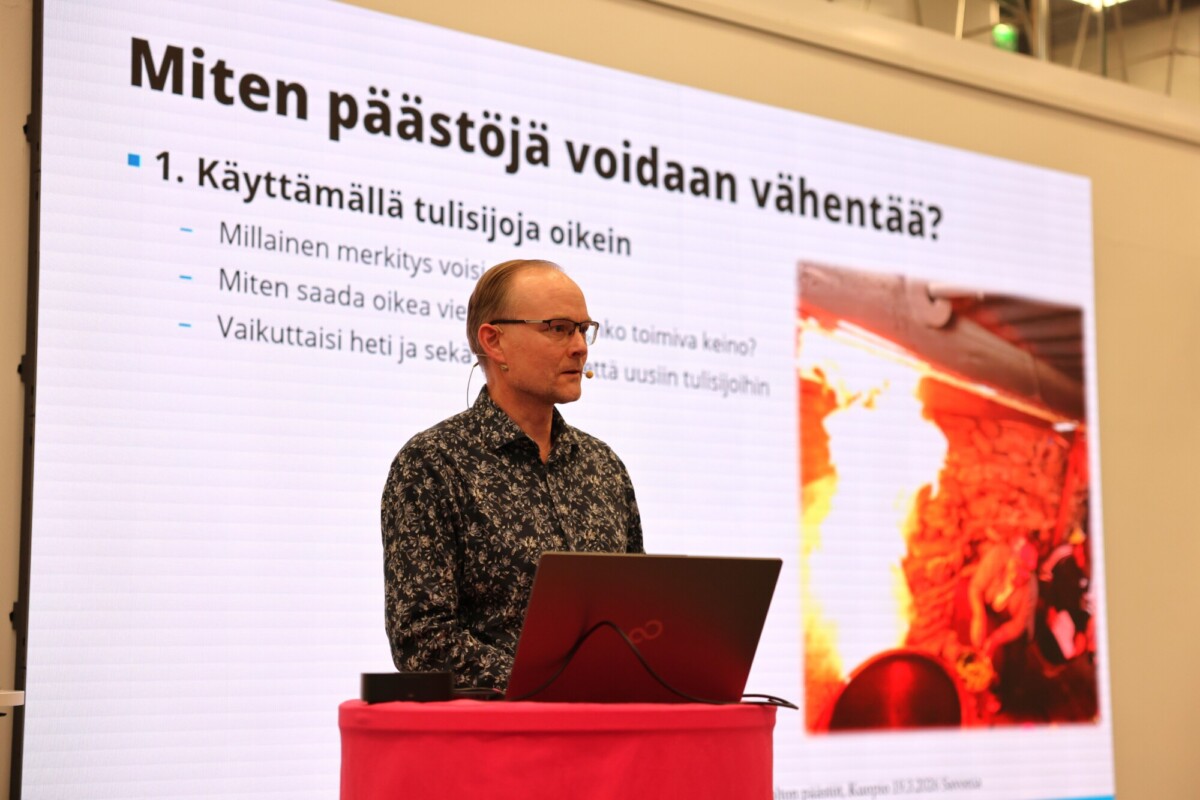 Mies seisoo korokkeella kannettavan tietokoneen kanssa ja puhuu suomeksi päästöjen vähentämisestä. Hänen takanaan olevalla suurella näytöllä näkyy pykäliä ja kuva takasta, jossa on liekkejä.