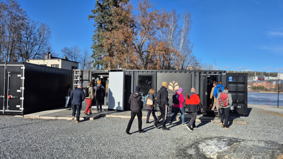 Ihmiset kävelevät sisään ja ulos mustista konttipohjaisista rakennuksista aurinkoisena päivänä, joillakin on takit yllään ja reput mukanaan. Taustalla on puita, joissa on harvat lehdet, ja järvi.