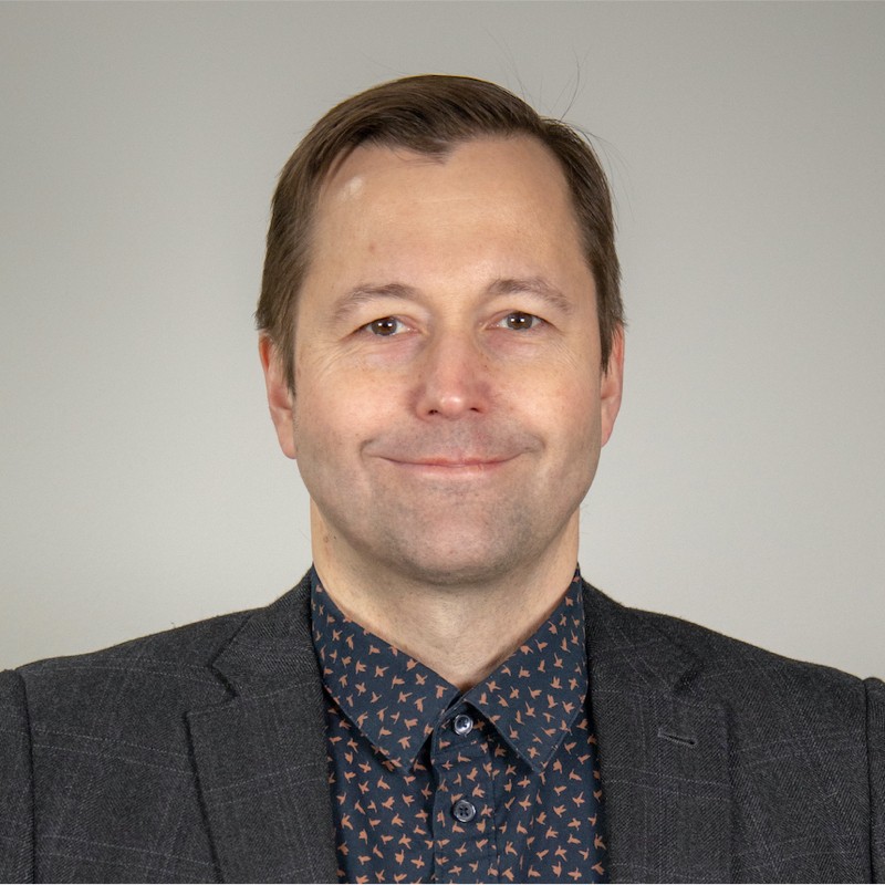 Lyhyet ruskeat hiukset omaava mies, jolla on tumma bleiseri ja tumma paita, jossa on pieni kuvio, hymyilee vaaleanharmaan taustan edessä.