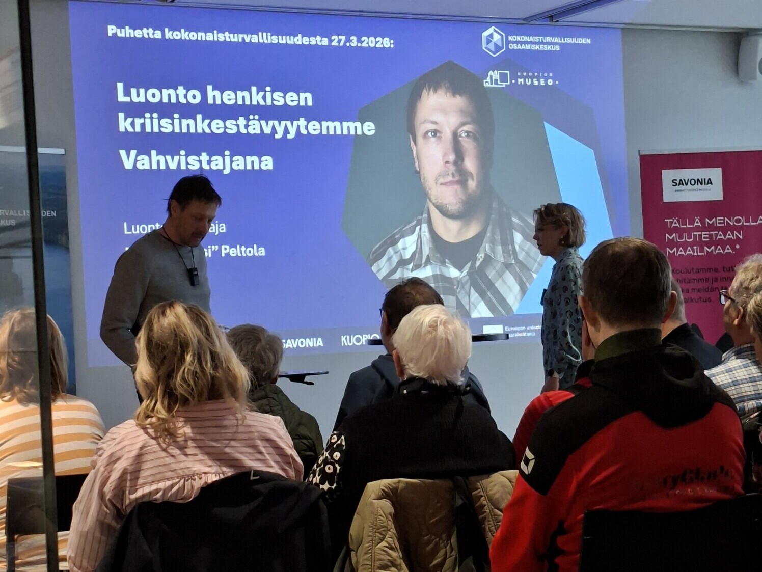 Ryhmä ihmisiä istuu suurta näyttöä vastapäätä, jossa näkyy esitys. Diassa näkyy miehen valokuva ja suomenkielistä tekstiä henkisestä kestävyydestä ja luonnosta. Mies ja nainen seisovat edessä valmistautumassa puhumaan.