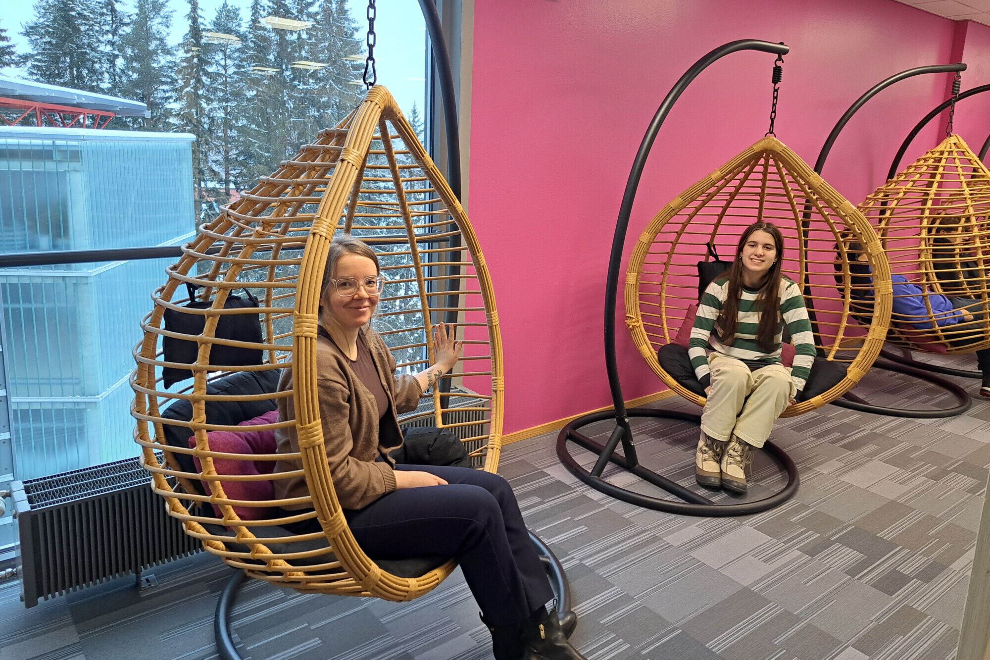 Kaksi naista istuu roikkuvissa punotuissa munatuoleissa kirkkaasti valaistussa huoneessa, jonka vaaleanpunaiset seinät ja suuret ikkunat näyttävät ulkona olevat lumiset puut. Heidän yläpuolellaan näkyy seinäkello. Naiset hymyilevät ja ovat rentoja.