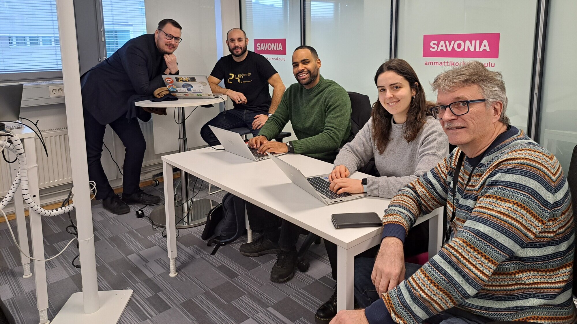 Modernissa toimistossa on viisi ihmistä, joista osa istuu pöydän ääressä kannettavien tietokoneiden kanssa ja osa seisoo. He hymyilevät kameraan päin. Taustalla näkyy kaksi julistetta, joissa on sana SAVONIA.