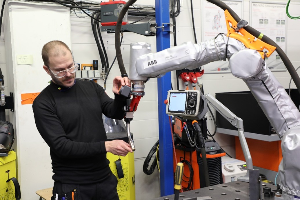 Suojalaseja käyttävä henkilö käyttää suurta robottikäsivartta teollisuusverstaassa säätämällä sen työkalua. Taustalla näkyy erilaisia laitteita, kaapeleita ja ohjauspaneeli.