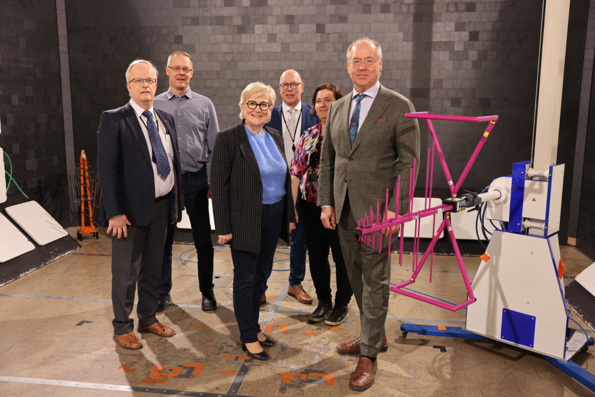 Kuusi ihmistä seisoo yhdessä teknisessä laboratorioympäristössä laitteiden ja antennien ympäröimänä. He ovat pukeutuneet työasuun ja poseeraavat ryhmäkuvassa hymyillen kameralle.