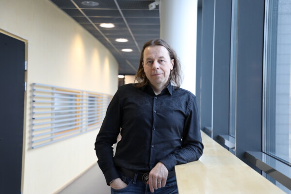 Mustaan paitaan ja farkkuihin pukeutunut pitkähiuksinen mies seisoo sisätiloissa suurten ikkunoiden äärellä nojaillen puiseen reunalistaan modernissa käytävässä, joka on täynnä luonnonvaloa.