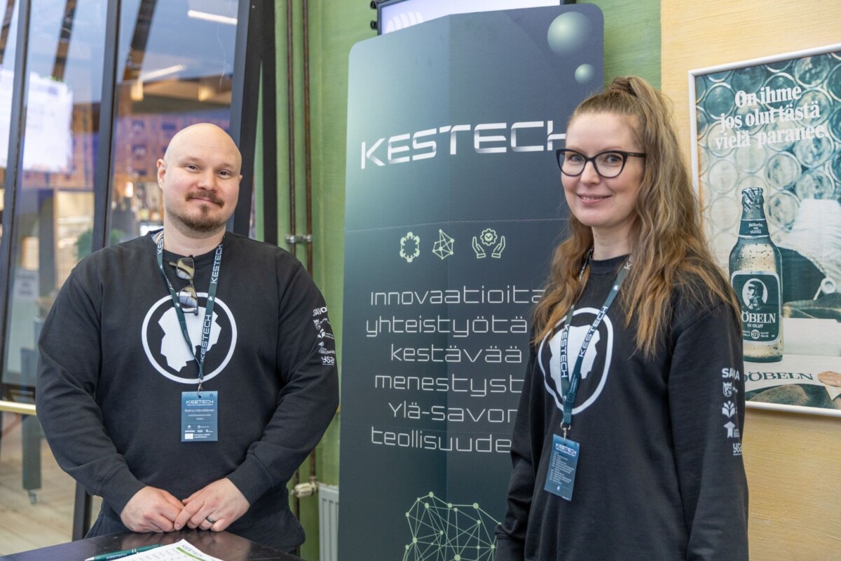 Kaksi ihmistä, joilla on samanlaiset tummat paidat ja kaulanauhat, seisovat bannerin vieressä, jossa on teksti KESTECH ja suomenkielisiä sanoja innovaatiosta ja yhteistyöstä sisätiloissa järjestettävässä tapahtumassa tai messuilla.