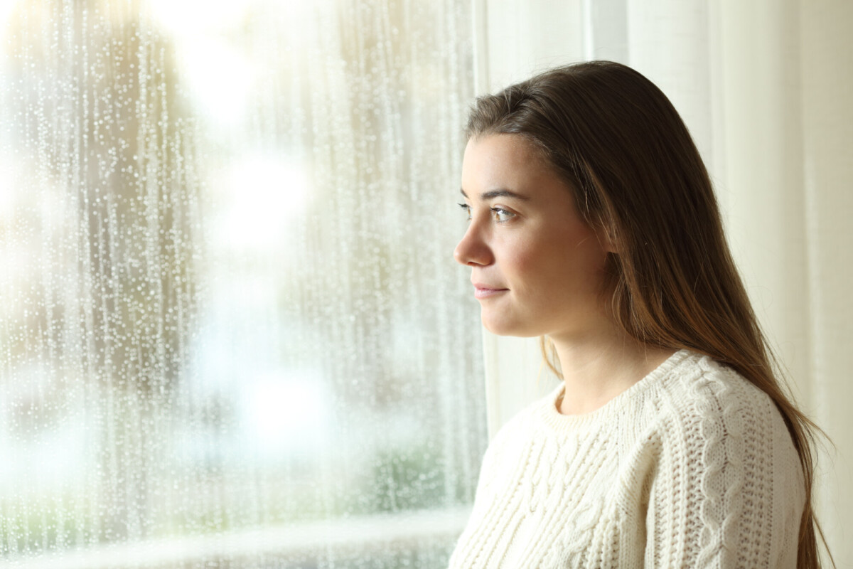 Nuori nainen, jolla on pitkät ruskeat hiukset ja valkoinen villapaita, katsoo mietteliäästi ulos ikkunasta sadepäivänä, jolloin lasissa näkyy sadepisaroita.
