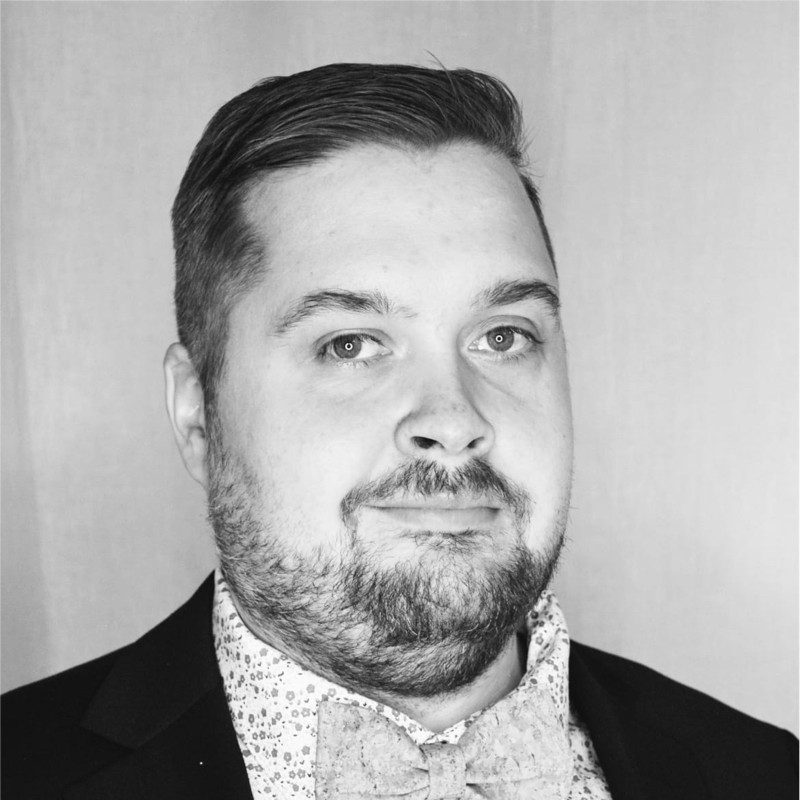 Mustavalkoinen muotokuva miehestä, jolla on lyhyet hiukset ja leikattu parta, päällään puvun takki, kuviollinen paita ja rusetti, katselee hieman sivulle neutraali ilme.