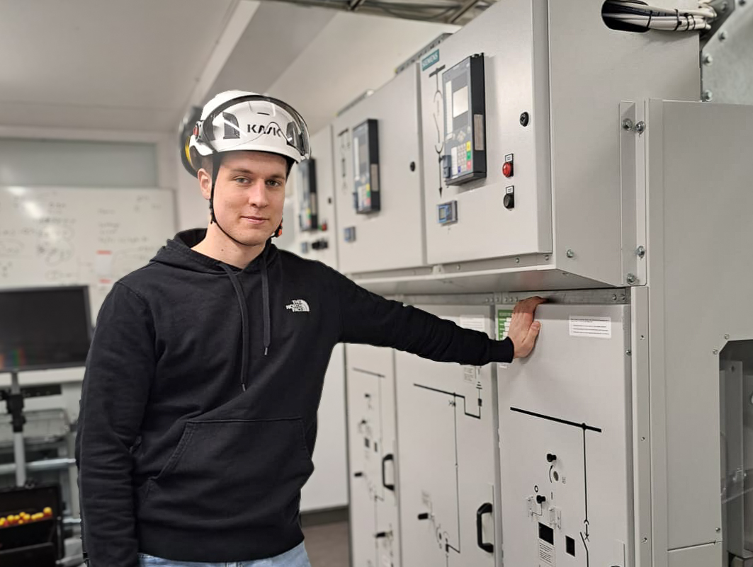 Valkoiseen suojakypärään ja mustaan huppariin pukeutunut mies seisoo teollisuuden valvontahuoneessa ja lepuuttaa kättään harmaalla sähköpaneelilla, jossa on erilaisia kytkimiä ja ilmaisimia.