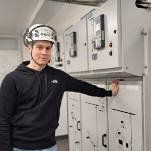 Valkoiseen suojakypärään ja mustaan huppariin pukeutunut mies seisoo teollisuuden valvomossa ja lepuuttaa kättään suurella sähkötaululla. Taustalla näkyy näyttöjä ja laitteita.