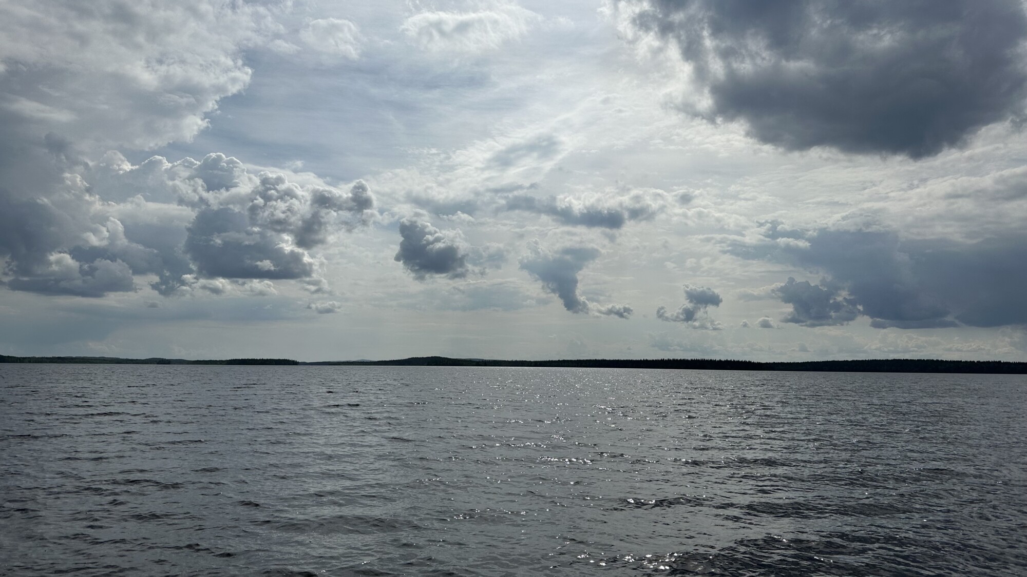 Suuri, rauhallinen järvi ulottuu horisonttiin asti enimmäkseen pilvisen taivaan alla. Auringonvalo kurkistaa pilvien läpi ja heijastuu veden aaltoilevasta pinnasta. Kaukana rannalla näkyy puuraja.