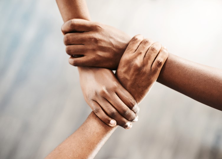 Neljä kättä tarttuu toistensa ranteisiin yhteen liitetyssä neliössä, joka symboloi yhtenäisyyttä, tiimityötä ja tukea, sumeaa neutraalia taustaa vasten.