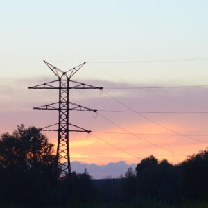 Voimajohdot ja siirtomastot ulottuvat puiden peittämään maisemaan, siluettina värikäs auringonlaskun taivas, jossa on oranssin, vaaleanpunaisen ja sinisen sävyjä.