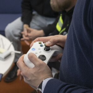 Henkilö pitelee valkoista videopeliohjainta, ja taustalla on muita henkilöitä, jotka istuvat lähellä, hieman epätarkasti. Asetelma vaikuttaa rennolta ja rennolta, ja näkyvissä on sininen sohva ja pöytä.