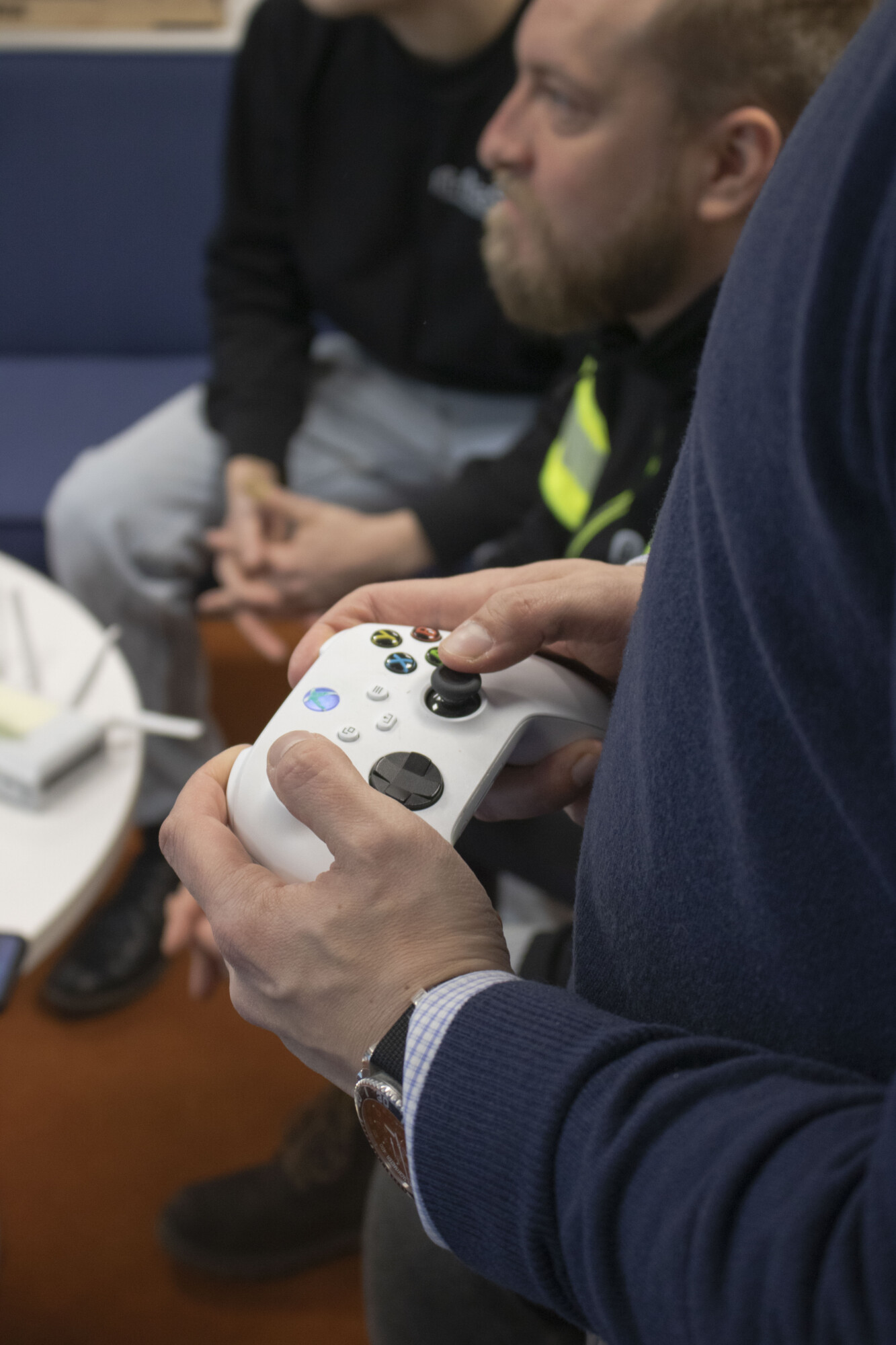 Henkilö pitelee valkoista videopeliohjainta, ja taustalla on muita henkilöitä, jotka istuvat lähellä, hieman epätarkasti. Asetelma vaikuttaa rennolta ja rennolta, ja näkyvissä on sininen sohva ja pöytä.