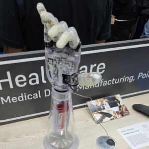 Robottikäsiproteesi on esillä näyttelyosastolla, ja taustalla on kyltti, jossa lukee osittain Healthcare Medical Device Manufacturing, Point of Care.