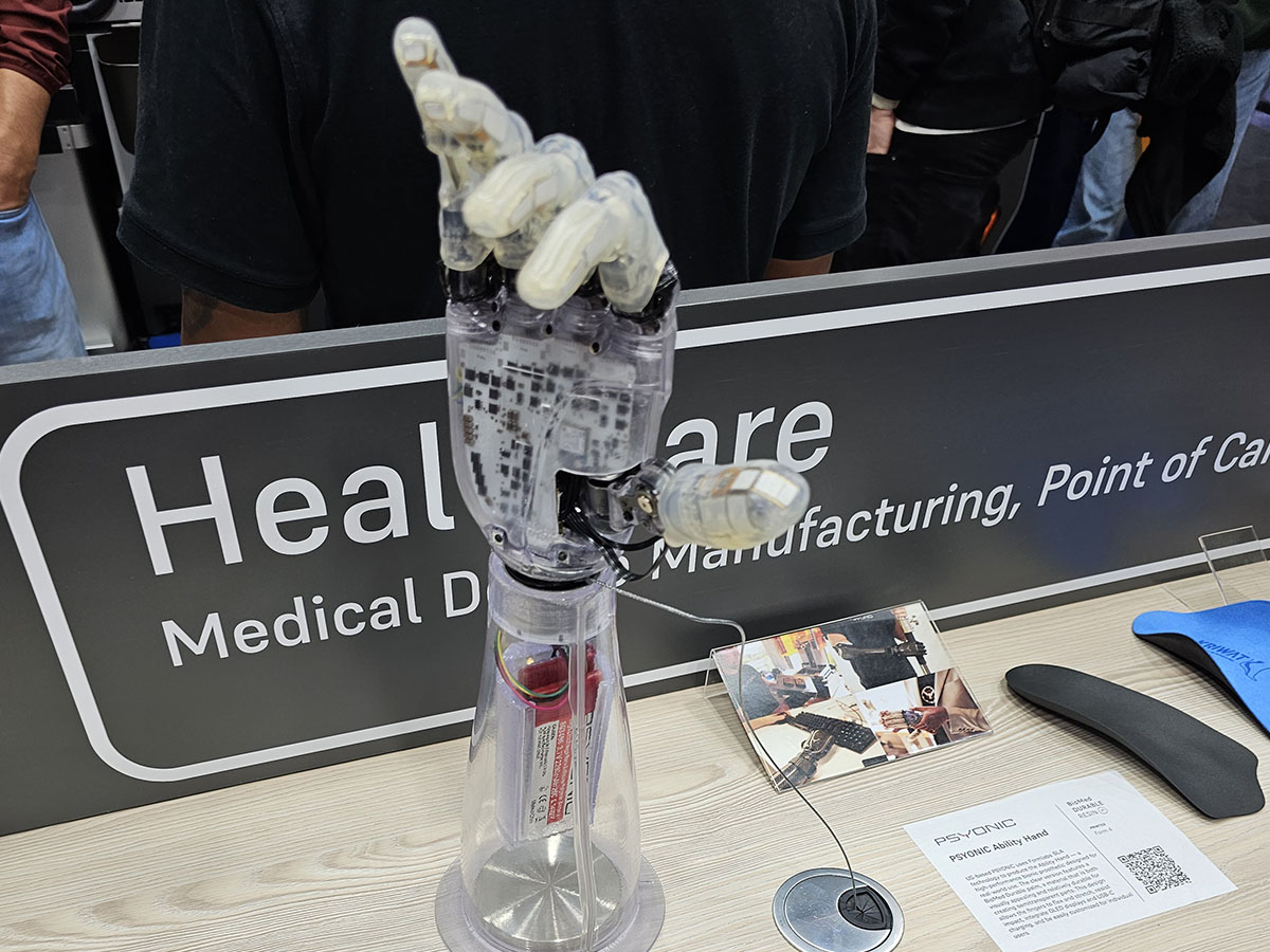 Robottikäsiproteesi on esillä näyttelyosastolla, ja taustalla on kyltti, jossa lukee osittain Healthcare Medical Device Manufacturing, Point of Care.