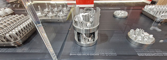 Mustalle pöytätasolle on sijoitettu erilaisia monimutkaisia metallisia 3D-tulostettuja osia ja komponentteja, jotka on sijoitettu pyöreille metallijalustoille ja jotka esittelevät kehittynyttä valmistustekniikkaa.