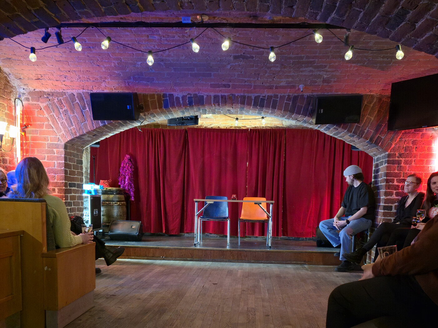 Pieni näyttämö, jossa on kaksi tuolia ja pöytä, on tiilikaarien alla, takana punaiset verhot. Yleisö istuu puisilla penkeillä ja tuoleilla odottamassa esitystä. Valonarut ja lämmin valaistus luovat viihtyisän tunnelman.