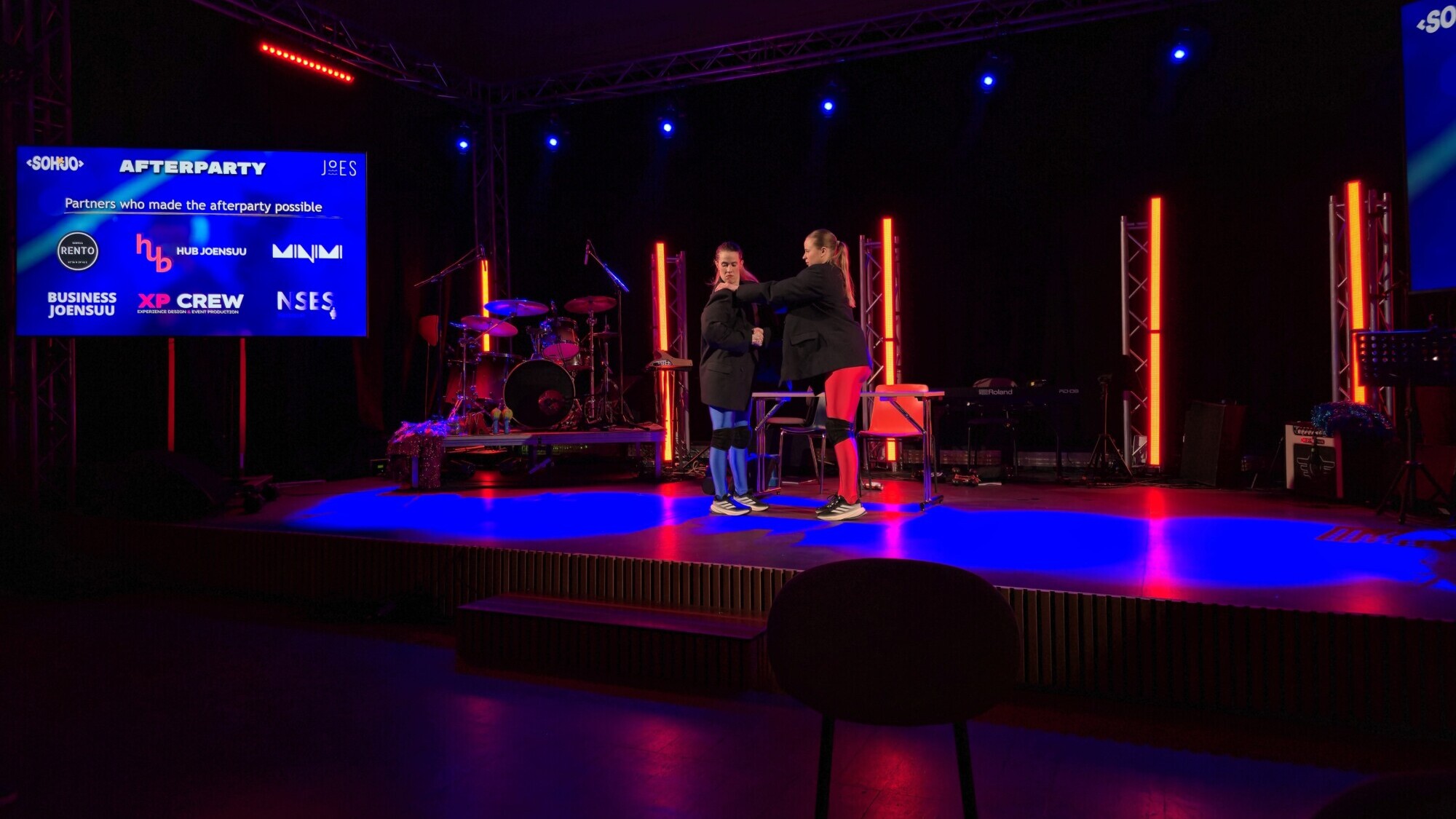 Kaksi ihmistä seisoo ja puhuu lavalla, jonka taustalla on värikäs valaistus ja soittimia. Suurella näytöllä näkyvät afterpartyjen sponsorit ja logot. Tapahtumapaikka on hämärästi valaistu, ja etualalla näkyy muutama tuoli.