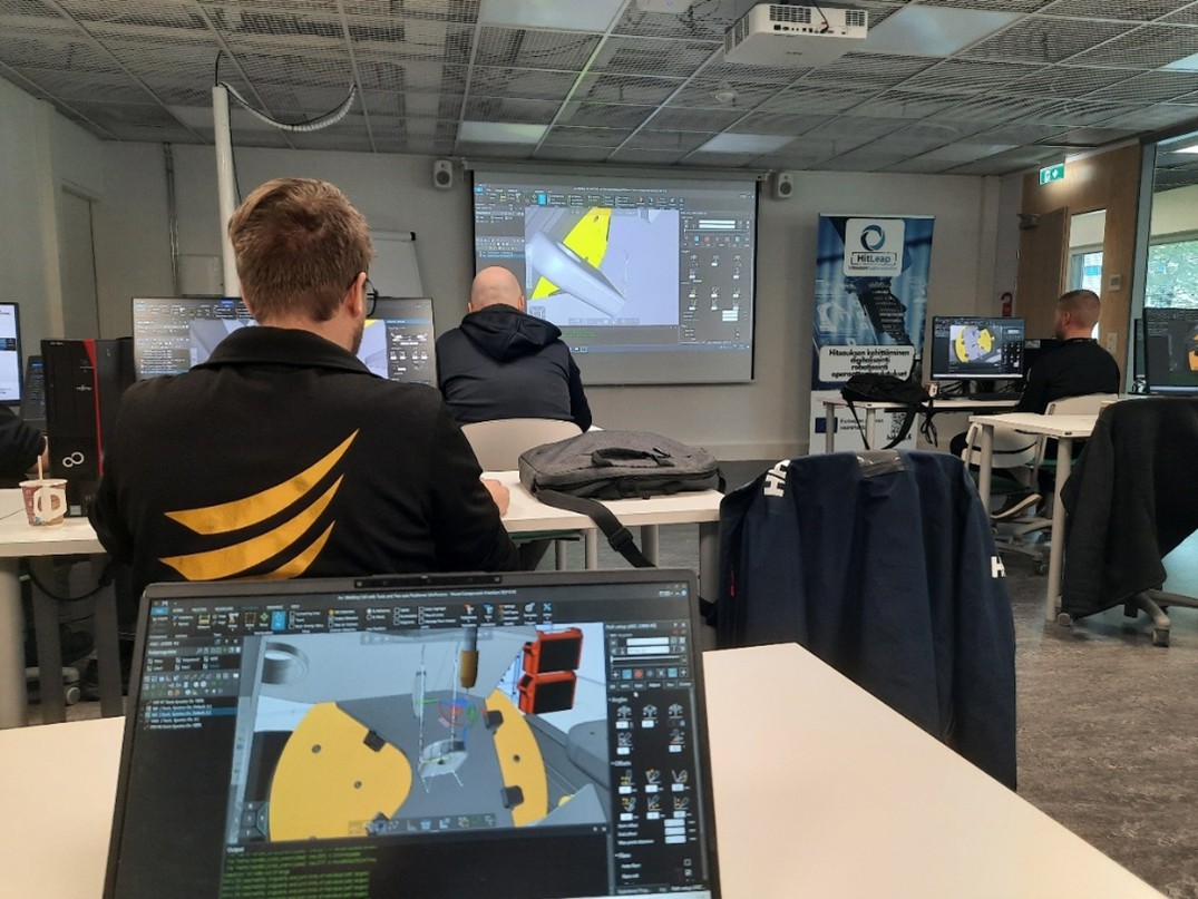 Ihmiset työskentelevät luokkahuoneessa tietokoneiden ääressä ja katselevat 3D-mallinnusohjelmia näytöillään. Opettaja esittelee vastaavaa ohjelmistoa projektorilla huoneen etuosassa. Näkyvissä on merkkitakkeja ja banderolleja.