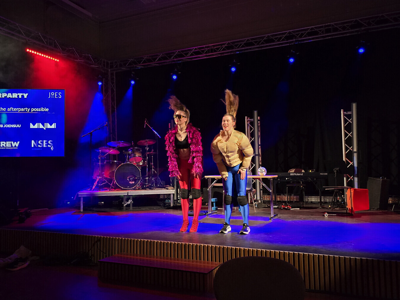 Kaksi esiintyjää seisoo lavalla värikkäiden valojen alla leikkisissä puvuissa: toinen on punapukuinen ja hänellä on suuret aurinkolasit, toinen on ruskehtava ja hänellä on korkea poninhäntä. Taustalla näkyy soittimia ja näyttö, jossa on tapahtuman tiedot.