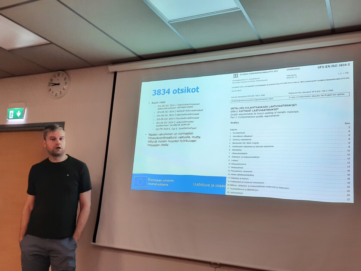 Mies seisoo valkokankaan vieressä, jossa näkyy esitysdia, jossa on suomenkielistä tekstiä, muun muassa otsikko 3834 otsikot ja luettelo standardeista ja aiheista. Huoneessa on valkoinen katto, kello ja poistumisopaste.