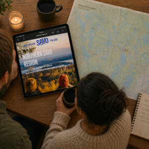 Kaksi ihmistä istuu pöydän ääressä kahvin ääressä ja katselee taulua, jossa on Pohjois-Savon maakunta ja Pohjois-Savon kartta. Pöydällä on myös muistilappu ja kynttilä.