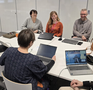 Seitsemän ihmistä istuu kokoushuoneessa pöydän ympärillä, ja jokaisella on kannettava tietokone tai muistikirja. He keskustelevat ja näyttävät hymyilevän ja nauttivan keskustelusta. Asetelma on rento ja yhteistoiminnallinen.