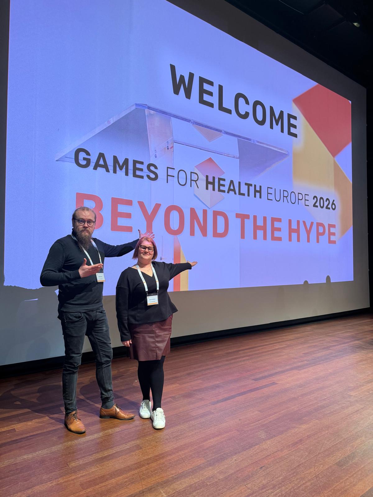 Kaksi ihmistä seisoo lavalla suuren näytön edessä, jossa lukee WELCOME. Games for Health Europe 2026. Hypen takana. Molemmat hymyilevät ja osoittavat elein kohti näyttöä.