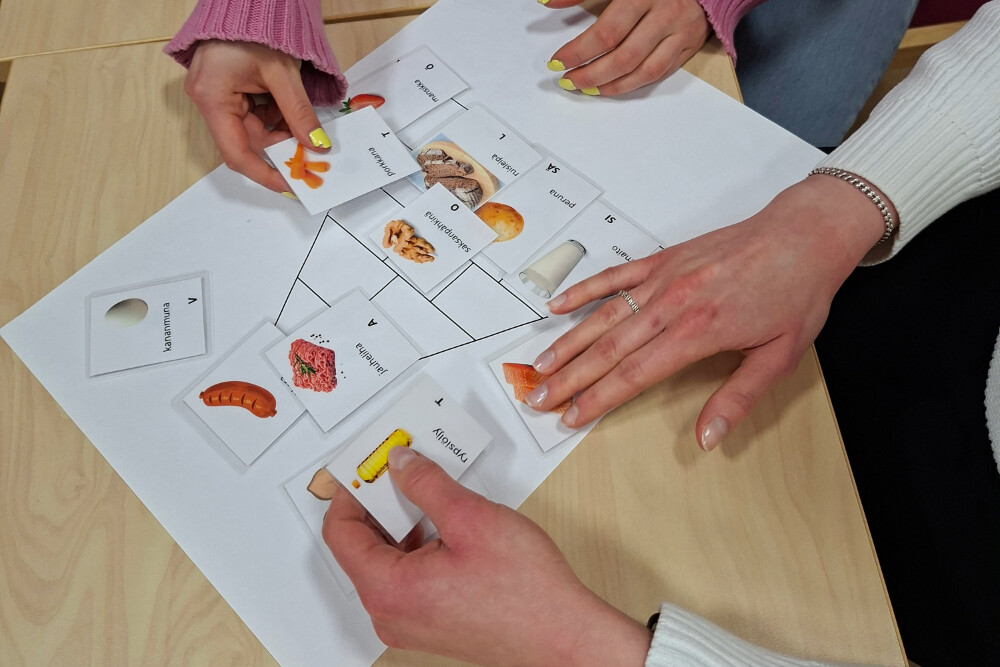 Kolme ihmistä järjestää kortteja, joissa on kuvia elintarvikkeista, kuten leivästä ja kalasta, ja tekstilappuja valkoiselle paperille, jossa on vuokaavio. Toiminta tapahtuu vaalealla puupöydällä.