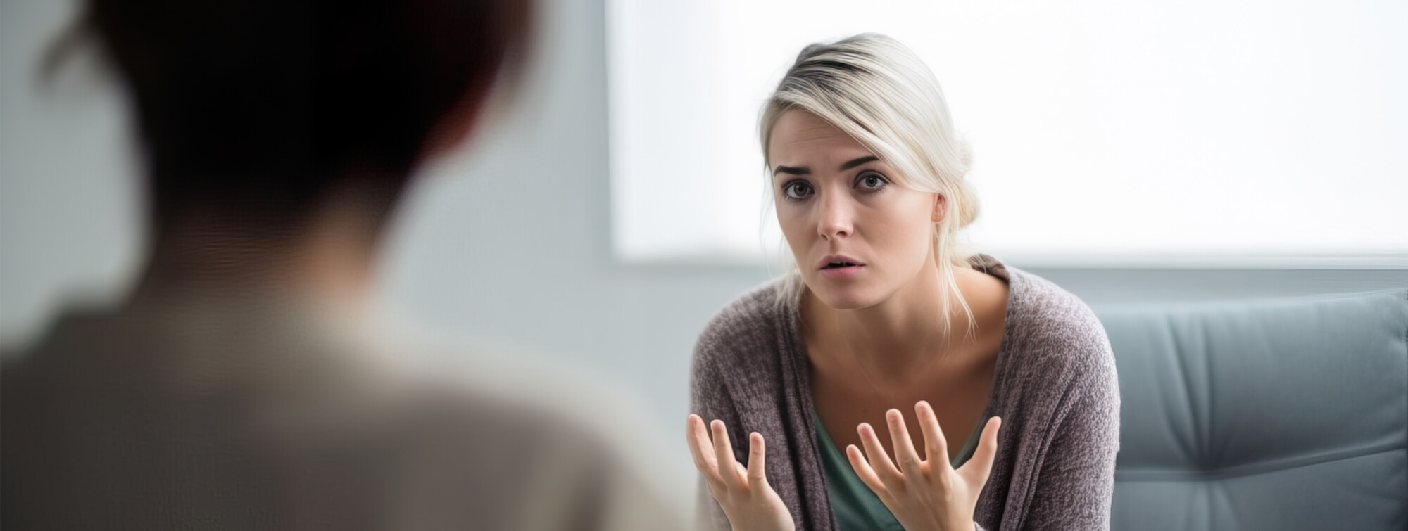 Vaaleahiuksinen huolestunut nainen istuu sisätiloissa, elehtii molemmilla käsillään ja puhuu toiselle henkilölle, jonka selkä näkyy etualalla. Tilanne vaikuttaa vakavalta keskustelulta tai neuvonpidolta.