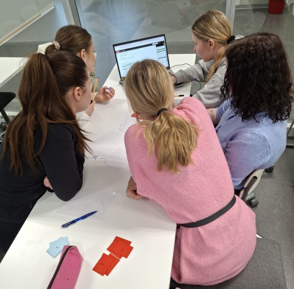 Neljä naista istuu valkoisen pöydän ympärillä, tekevät yhteistyötä paperitöiden parissa ja katselevat kannettavan tietokoneen näyttöä. Kynät, paperit ja värilliset kortit ovat hajallaan pöydällä valoisassa, modernissa huoneessa.