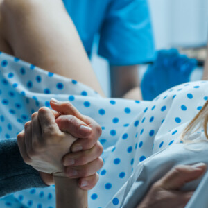 Sairaalamekkoon pukeutunut nainen synnyttää ja tarttuu jonkun käteen tukena, kun hoitohenkilökunta avustaa häntä synnytyksen aikana.