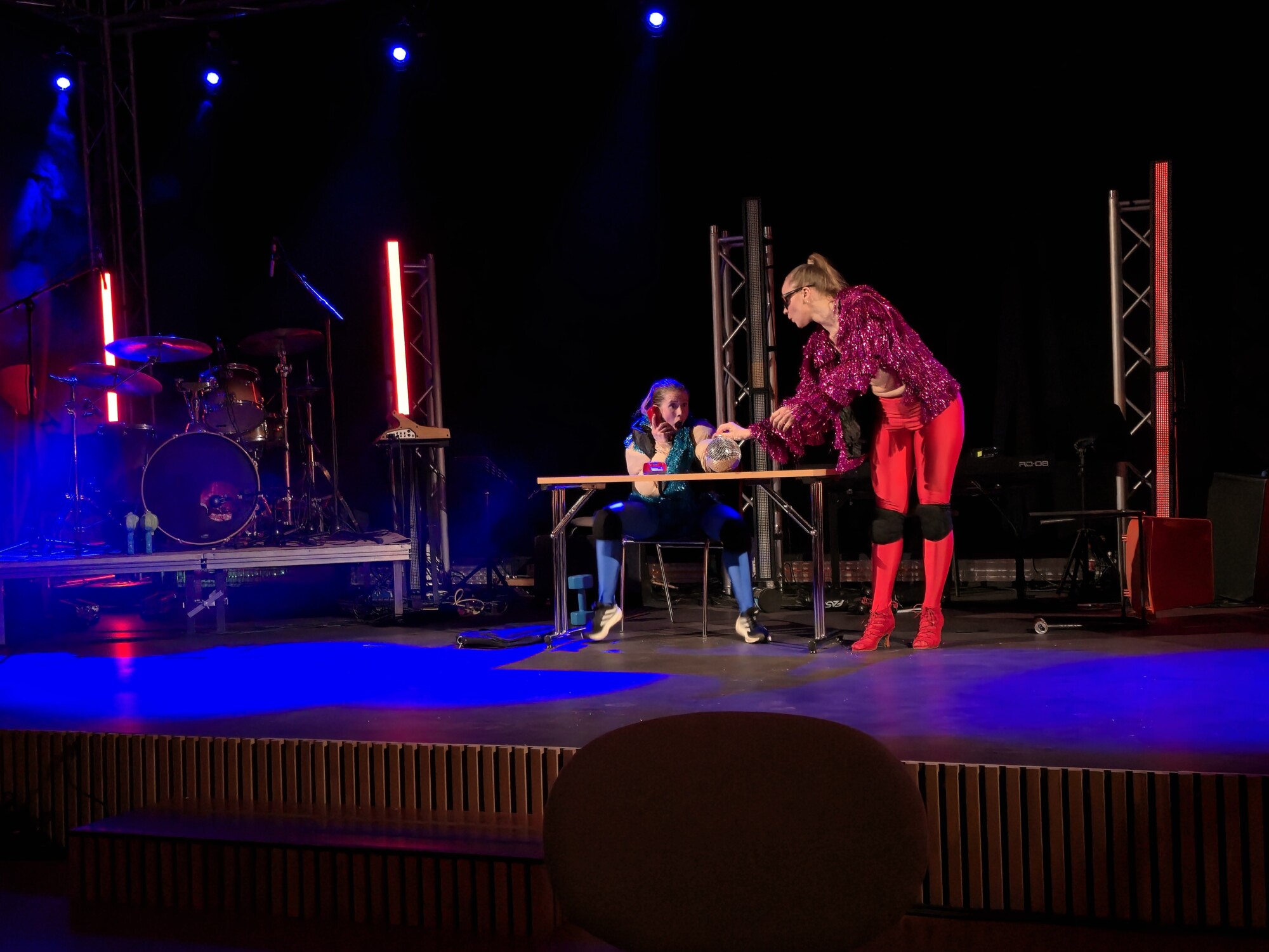 Kaksi värikkäisiin pukuihin pukeutunutta esiintyjää on lavalla pöydän ääressä. Toinen istuu ja toinen seisoo ja ojentaa valkoista esinettä. Taustalla on näyttämövaloja ja soittimia.