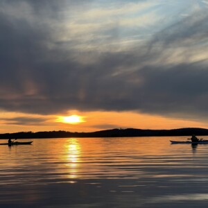 Kaksi kajakissa olevaa henkilöä meloo rauhallisella vedellä auringonlaskun aikaan, kun järvellä heijastuvat dramaattiset pilvet ja kultainen auringonvalo. Taustalla näkyy siluettimaisia kukkuloita.