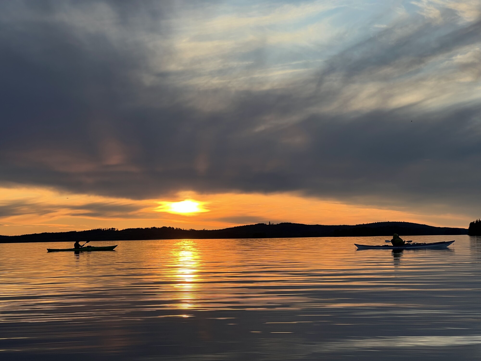 Kaksi kajakissa olevaa henkilöä meloo rauhallisella vedellä auringonlaskun aikaan, kun järvellä heijastuvat dramaattiset pilvet ja kultainen auringonvalo. Taustalla näkyy siluettimaisia kukkuloita.