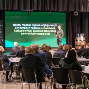 Puhuja seisoo lavalla esittelemässä puheenvuoroaan pöydissä istuvalle yleisölle. Hänen takanaan olevalla suurella näytöllä näkyy suomenkielistä tekstiä tekoälystä ja digitaalisista järjestelmistä. Ympäristö on sisätiloissa järjestettävä tapahtuma tai konferenssi.