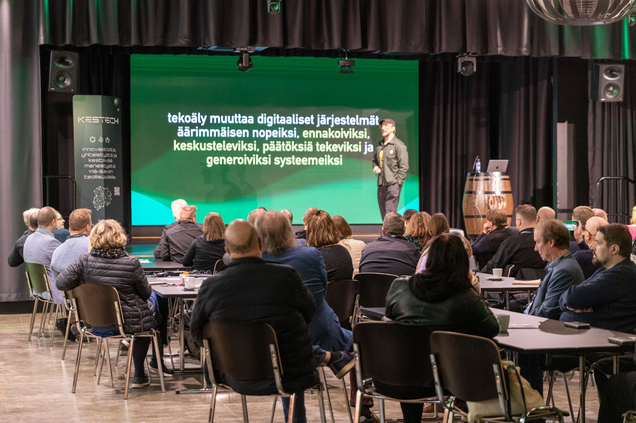 Puhuja seisoo lavalla esittelemässä puheenvuoroaan pöydissä istuvalle yleisölle. Hänen takanaan olevalla suurella näytöllä näkyy suomenkielistä tekstiä tekoälystä ja digitaalisista järjestelmistä. Ympäristö on sisätiloissa järjestettävä tapahtuma tai konferenssi.