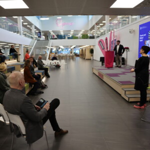 Nainen puhuu istuvalle yleisölle modernissa, avoimessa tilassa, jossa on porrastetut istuimet. Hänen takanaan mies seisoo suuren digitaalisen näytön äärellä, jossa näkyy kuvia ja tekstiä. Ympäristö on valoisa ja avara.