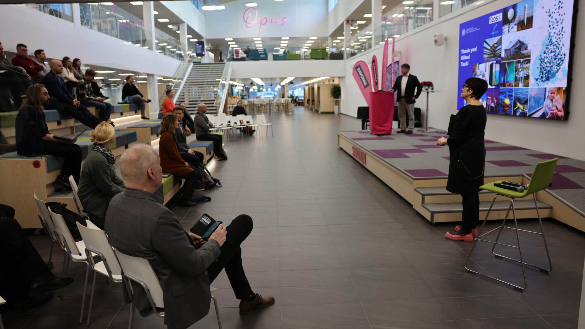 Nainen puhuu istuvalle yleisölle modernissa, avoimessa tilassa, jossa on porrastetut istuimet. Hänen takanaan mies seisoo suuren digitaalisen näytön äärellä, jossa näkyy kuvia ja tekstiä. Ympäristö on valoisa ja avara.