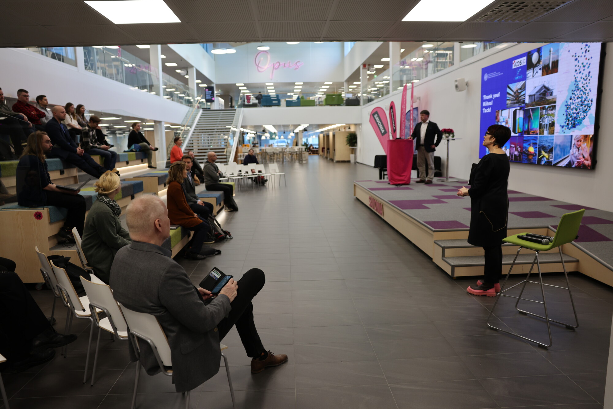 Nainen puhuu istuvalle yleisölle modernissa, avoimessa tilassa, jossa on porrastetut istuimet. Hänen takanaan mies seisoo suuren digitaalisen näytön äärellä, jossa näkyy kuvia ja tekstiä. Ympäristö on valoisa ja avara.