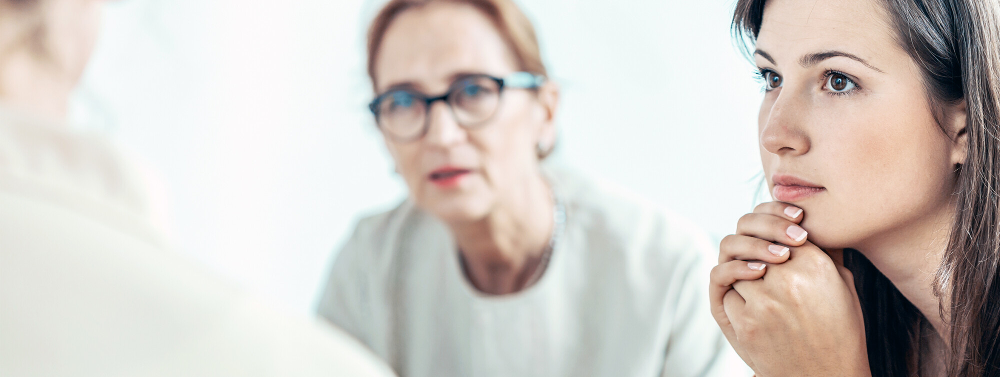 Nuori nainen istuu kädet leuan alla ja katsoo mietteliäästi. Taustalla on vanhempi silmälasipäinen nainen, joka puhuu hänelle, molemmat valoisassa, heikosti valaistussa huoneessa.