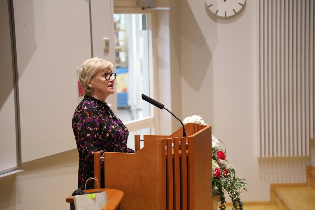 Lyhyet vaaleat hiukset ja silmälasit päässä oleva nainen seisoo puisella korokkeella ja puhuu mikrofoniin hyvin valaistussa huoneessa. Korokkeella on kukkia ja hänen takanaan seinällä on suuri kello.
